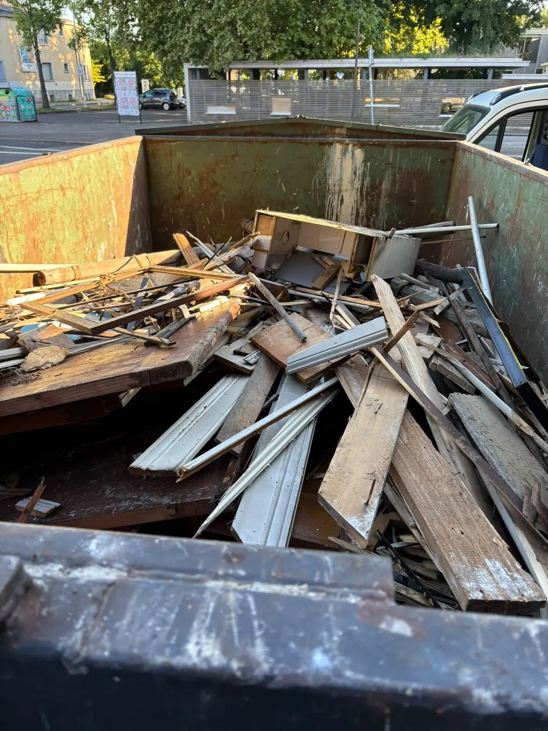 Benne remplie de bois de démolition, plinthes et chambranles, sur un parking urbain
