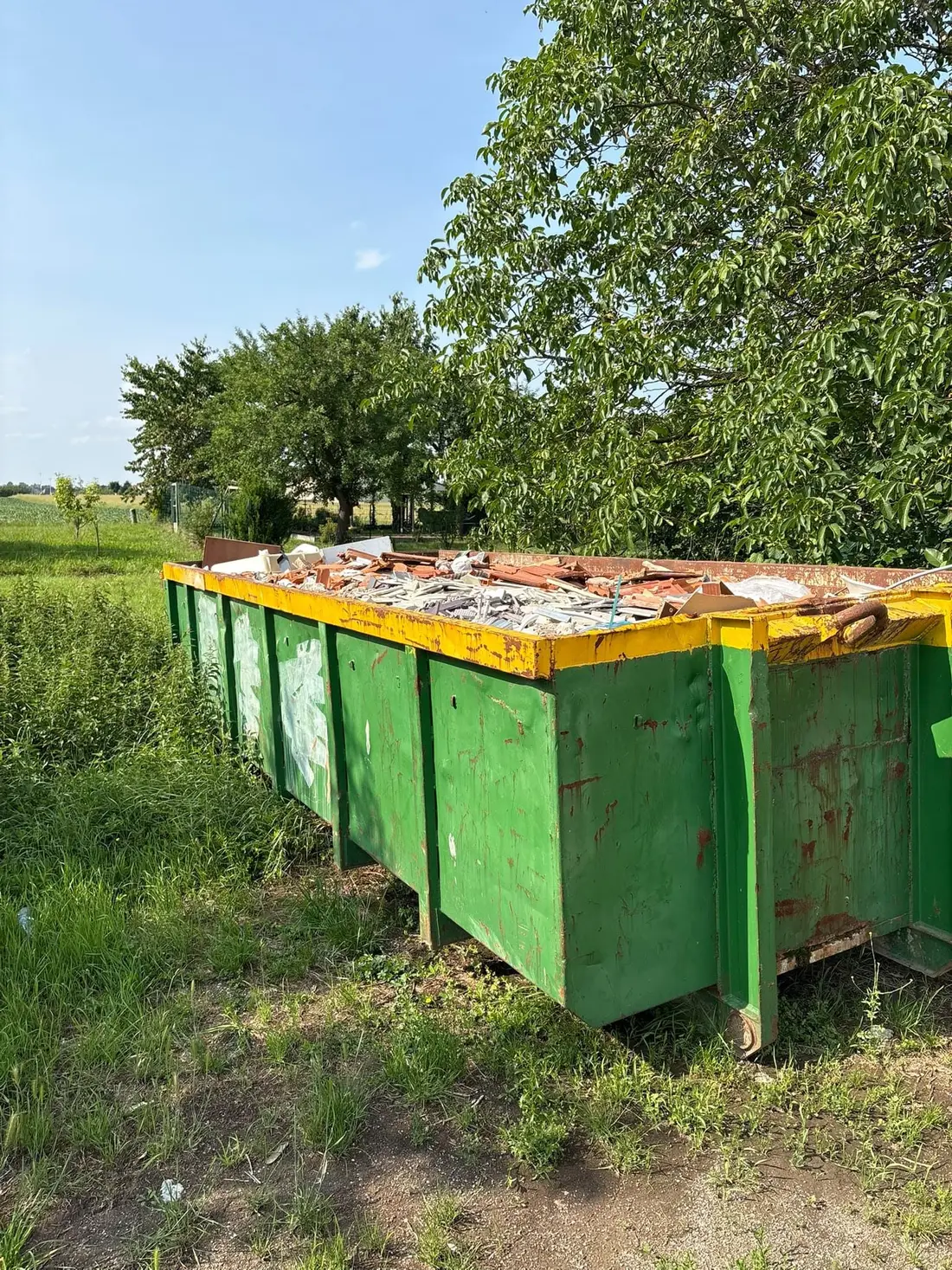 Grande benne remplie de gravats et de tuiles, posée en bordure de champ