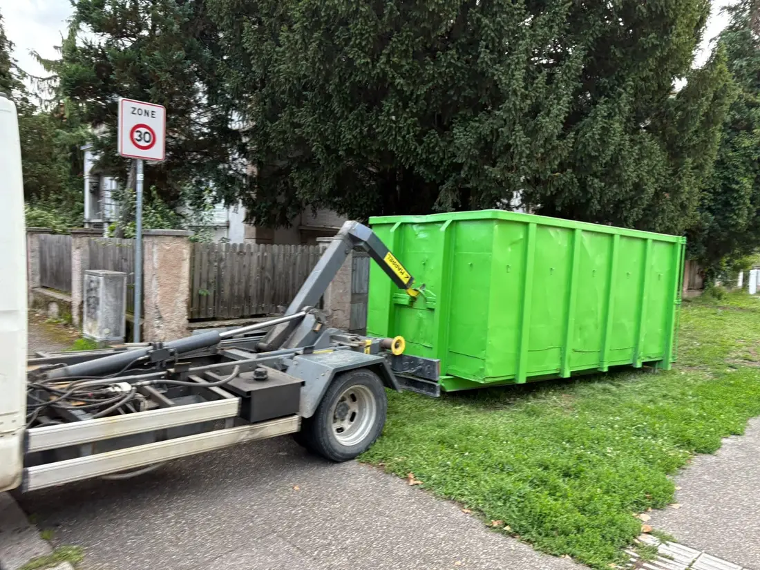 Camion ampliroll déposant une benne verte de 20 m³ sur un terrain enherbé, dans une rue en zone 30